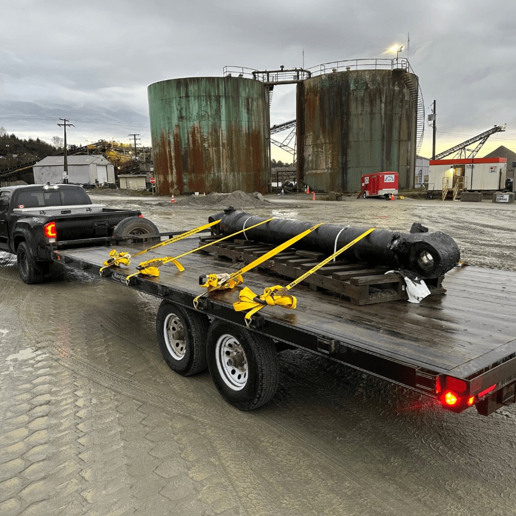 Black Toyota Tacoma towing a flat deck trailer with pipe that is strapped down with yellow safety trailer straps, in Kitimat, BC.
